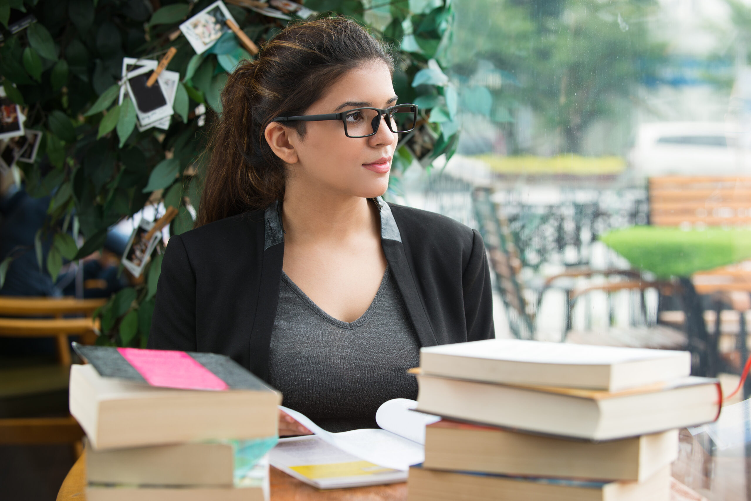 Portrait of pensive young Caucasian female student or teacher wearing glasses sitting at table with books and looking away