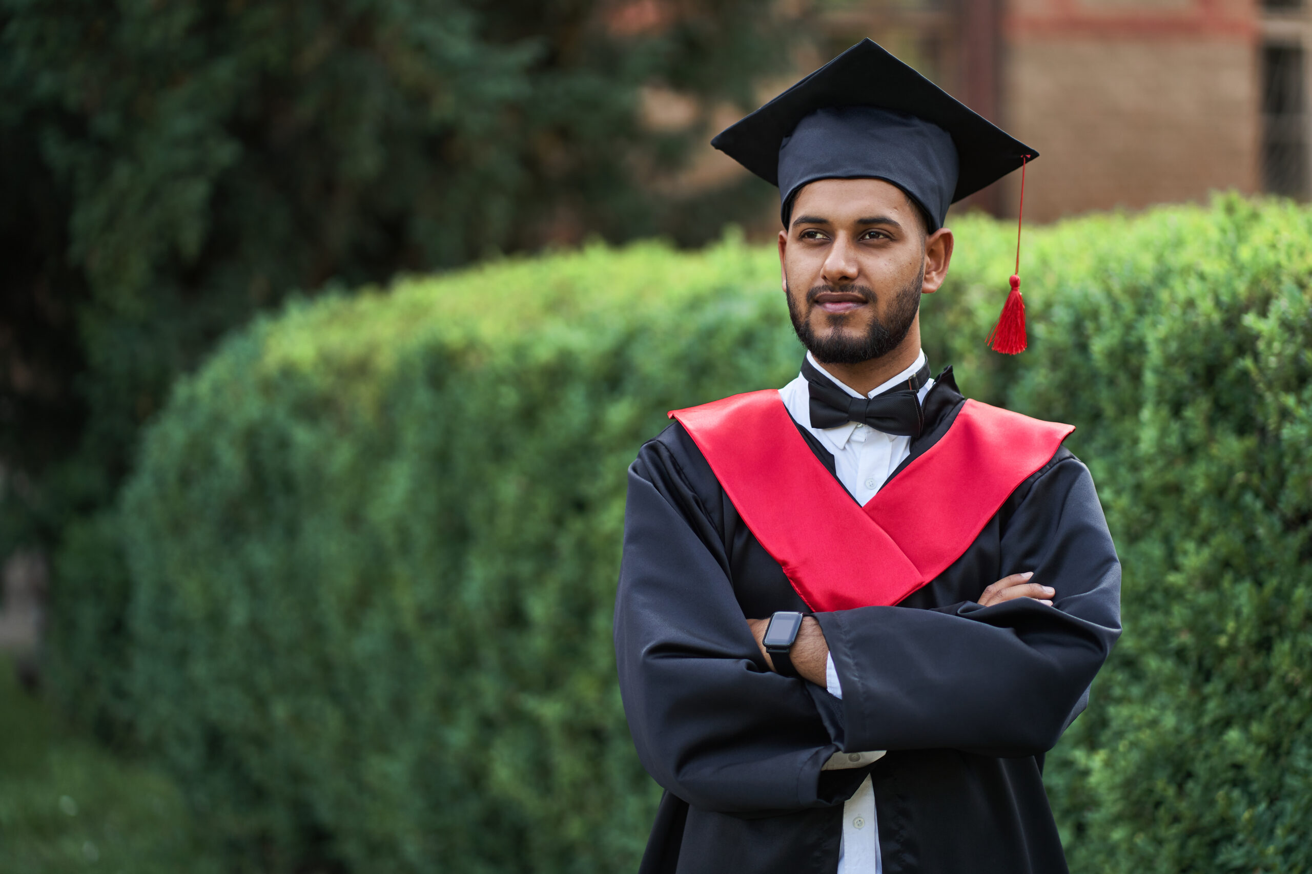 Serious indian graduate in graduation robe with crossed arms looking forward, copy space for text.