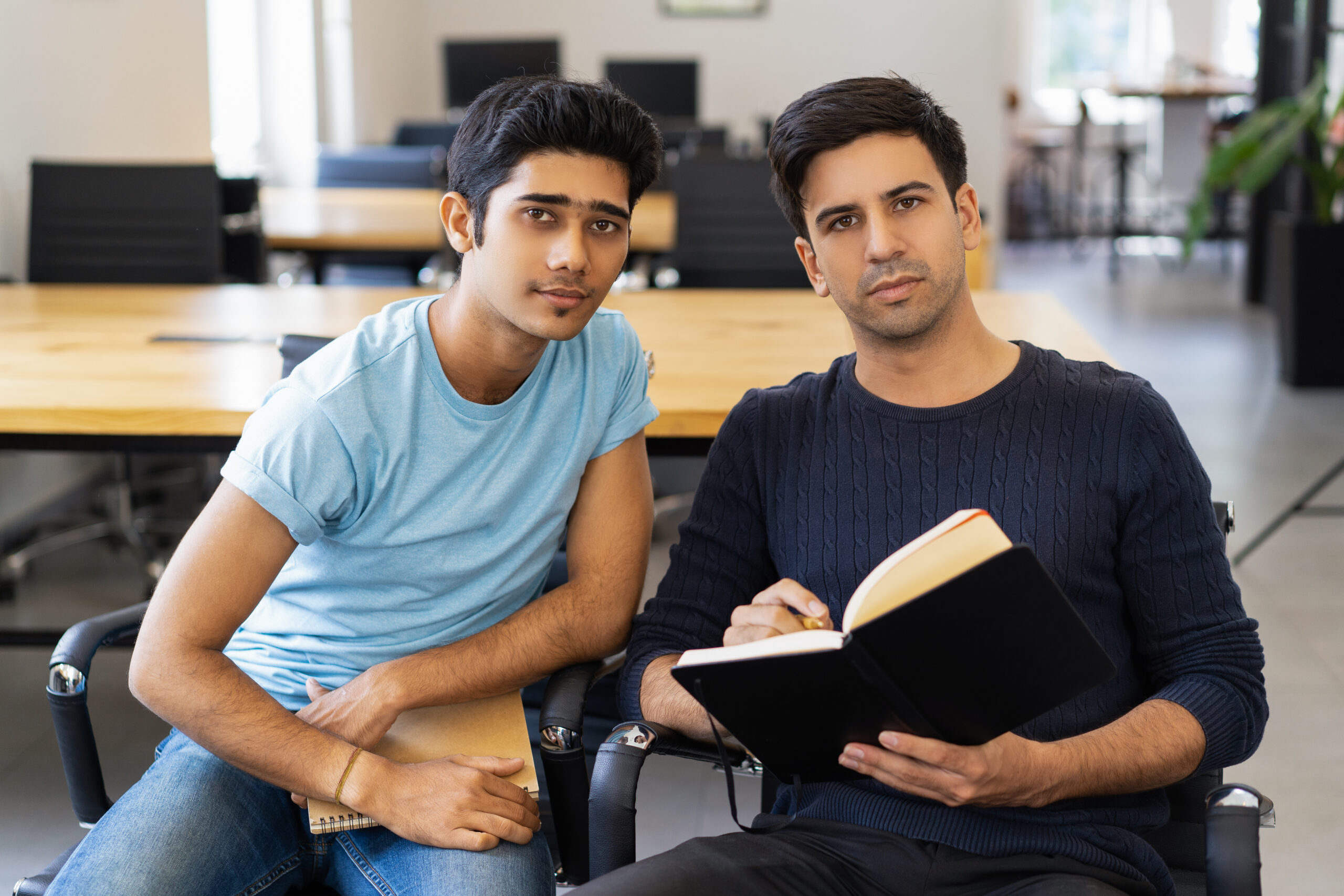 Two serious fellow students studying and looking at camera. Young men sitting in armchairs in classroom or library. Education and friendship concept. Front view.